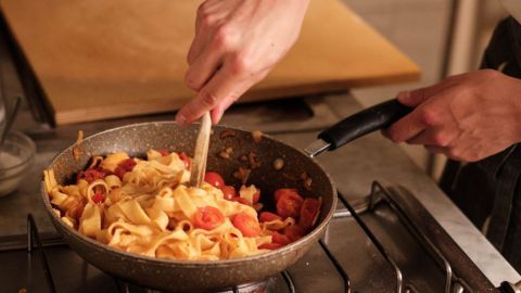 Eine Hand rührt in einer Pfanne mit Pasta und Tomaten auf einem Herd in einer Küche, zeigt italienische Kochkunst.