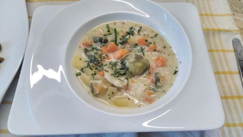 Eine Gemüsesuppe in einem weißen Teller zeigt viele bunte Gemüsewürfel und Kräuter, serviert auf einem karierten Tuch.