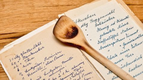 Ein Holzlöffel liegt auf handgeschriebenen Rezepten, darunter „Blattnockerln“, auf einem Holztisch.