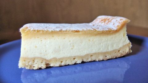 Superlockerer Käsekuchen: Geschlagene Sahne vor dem Backen unter die Käsemasse heben. Ein Käsekuchenstück liegt auf einem blauen Teller, leicht bestäubt mit Puderzucker, locker und cremig in der Textur.