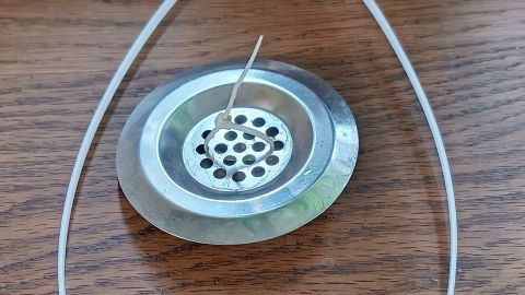 Ein metallisches Abflusssieb liegt auf einem Holztisch, fixiert mit einem Kabelbinder. Ein Zahnstocher steckt in den Löchern. Text: 17. Sept. 2024, 13:28.
