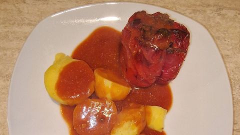 Eine gefüllte Paprika liegt auf einem weißen Teller, umgeben von Kartoffeln und einer Tomatensoße. Der Teller liegt auf einem hellen Holztisch.