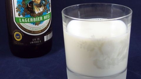 Ein Glas mit weißer, schaumiger Flüssigkeit steht neben einer Bierflasche, die im Hintergrund platziert ist.
