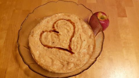 Bratapfelmus mit Marzipan in einer Schüssel, dekoriert mit einem Herz aus Sauce; ein Apfel liegt daneben auf dem Holztisch.