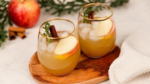 Genieße den weihnachtlichen Apfel-Zimt-Spritz: Ein erfrischender Cocktail mit Apfelscheiben, Zimt und frischem Thymian auf einem Holzbrett serviert.