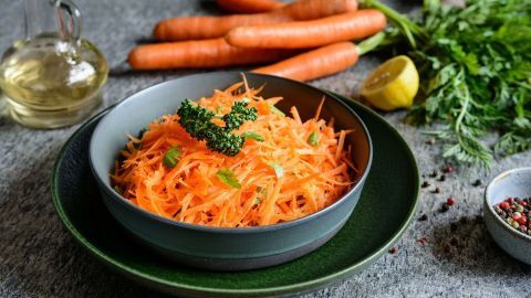 Rasierter Karottensalat liegt in einer Schüssel, während frische Karotten und Kräuter im Hintergrund stehen.