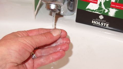 Eine Hand hält Wasser unter einem Wasserhahn, während im Hintergrund eine Verpackung mit Natron sichtbar ist.