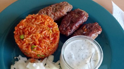 Genieße selbstgemachte Cevapcici mit würzigem Djuvec-Reis, frischen Zwiebeln und cremigem Dip: ein schmackhaftes Balkan-Erlebnis.
