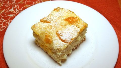 Ein Stück schneller Obstkuchen liegt auf einem weißen Teller. Es zeigte sich eine luftige Textur, bestreut mit Puderzucker und fruchtigen Stücken. Der Teller steht auf einem dekorativen, orangenen Untergrund.