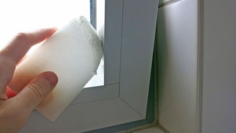 Ein Radierschwamm wird verwendet, um einen Fensterrahmen zu reinigen, während sich im Hintergrund helle Wände und Fliesen befinden.