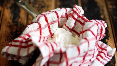 Ein Tuch mit Rot-weißem Karomuster umhüllt frisch zubereiteten Frischkäse, der auf einer rustikalen Holzoberfläche liegt.