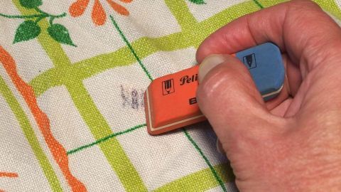 Eine Hand benutzt einen Radiergummi, um einen Kugelschreiberfleck von einem bunten Tischtuch zu entfernen.