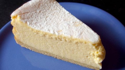 Ein Stück Käsekuchen "Ruck Zuck" liegt auf einem blauen Teller und sieht cremig und leicht pudrig aus.