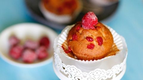 Ein Himbeer-Muffin steht auf einem eleganten weißen Ständer, umgeben von weiteren Muffins und frischen Himbeeren.