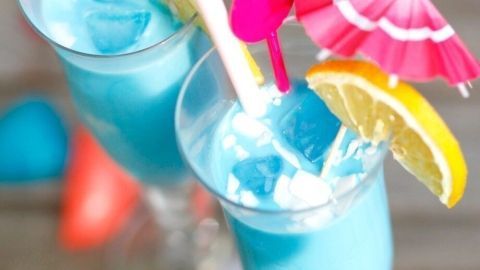 Zwei bunte Cocktails mit blauer Flüssigkeit, dekoriert mit kleinen Schirmen und Zitronenscheiben, stehen auf einem Tisch am Pool.