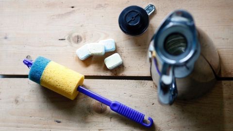 Eine Thermoskanne steht auf einem Tisch, zusammen mit einem Reinigungsschwamm und Bröckchen, um Tee- und Kaffeereste zu entfernen.