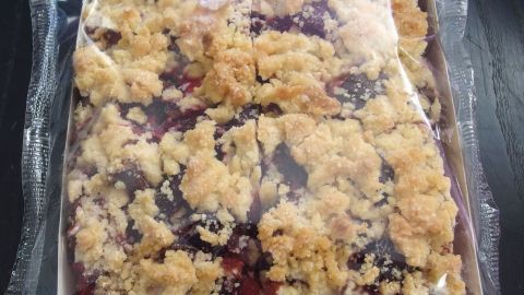 Ein tiefgekühlter Kuchen mit knusprigem Belag liegt in einer transparenten Verpackung auf einem dunklen Tisch.
