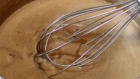 Eine silberne Küchenhilfe rührt in einer flüssigen Soße, die in einem metallenen Kochtopf auf dem Herd köchelt.