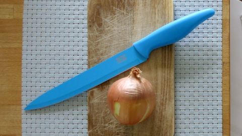Dieser Tipp erspart Taucherbrillen, Kontaktlinsen und vor allem Tränen, beim Zwiebel schneiden. Eine Zwiebel liegt auf einem Holzbrett, während ein türkiser Messer daneben bereit zum Schneiden ist.