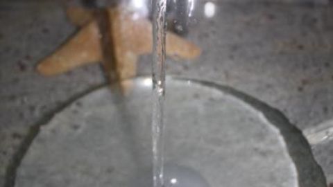 Wasser fließt aus einem Wasserhahn in ein gläsernes Waschbecken, dahinter liegt ein Seestern auf einem steinernen Untergrund.