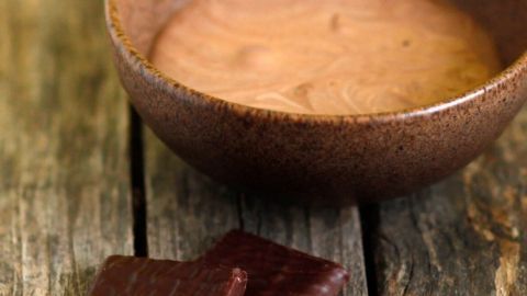 Eine Holzschale mit Pfefferminz-Schokoladentäfelchen-Creme steht auf einem rustikalen Holztisch, daneben liegen zwei Schokoladentäfelchen.