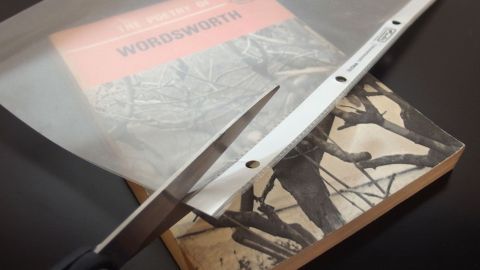 Eine Schere schneidet durch eine transparente Bucheinbandfolie, die auf einem Buch liegt. Der Hintergrund ist dunkel.