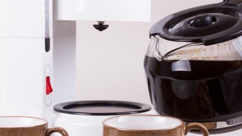 Eine Kaffeekanne wird über zwei Tassen gegossen, während im Hintergrund eine Kaffeemaschine steht.