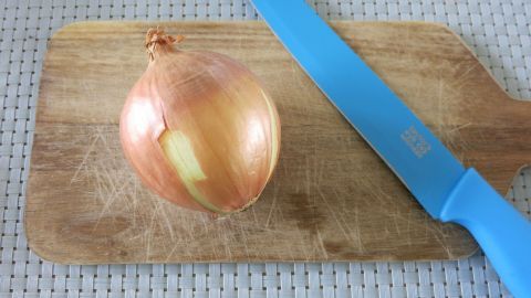 Hilfe bei sehr leichten Verbrennungen: Eine kleine Zwiebel halbieren und die frisch aufgeschnittene Seite der Zwiebel auf die verbrannte Stelle der Haut drücken. Die Entstehung einer Brandblase wird zwar nicht verhindert, aber die Schmerzen gelindert. Auf einem Holzbrett liegt eine Zwiebel mit einer blauen Klinge daneben, die bereit ist, zur Linderung leichter Verbrennungen verwendet zu werden.