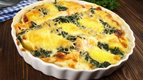 Ein Spinat-Lachs-Auflauf ist goldbraun überbacken und steht frisch auf einem Tisch, umgeben von Kräutern und einer blauen Serviette.