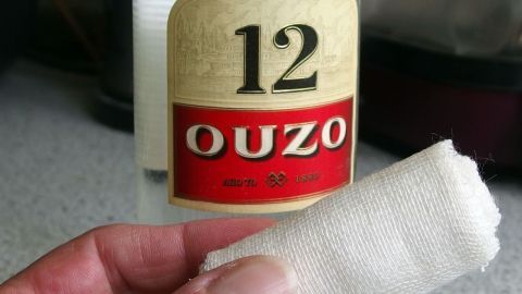 Eine Hand hält ein Tuch neben einer Flasche Ouzo, möglicherweise zur Linderung von Halsweh in einer küchenähnlichen Umgebung.