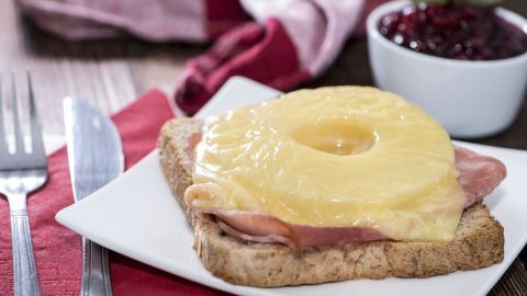 Ein Hawaiitoast liegt auf einem weißen Teller, belegt mit Schinken, Ananas und Käse, umgeben von Besteck und einem Schälchen Marmelade.