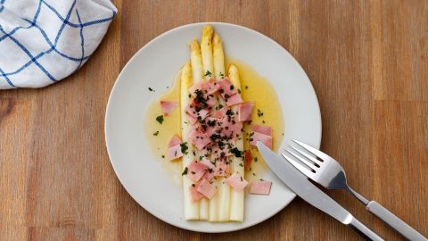 Weißer Spargel liegt auf einem Teller, umgeben von geschmolzener Butter und Schinkenstücken, auf einem Holztisch.