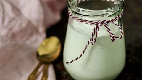 Ein Glas mit cremigem Joghurtgetränk steht auf einer rustikalen Oberfläche, begleitet von einem goldenen Löffel und Tuch.