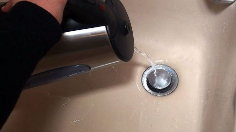 Eine Hand gießt Wasser aus einem Wasserkocher in einen Abfluss, um ihn mit Spülmittel zu reinigen.