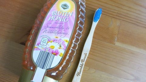 Bester Tipp gegen spröde Lippen: Lippen mit einer Zahnbürste und Melkfett abrubbeln und als Abschluss Honig auftragen. Ein Honigflasche und eine Zahnbürste liegen auf einem Holztisch, bereit zur Pflege spröder Lippen.