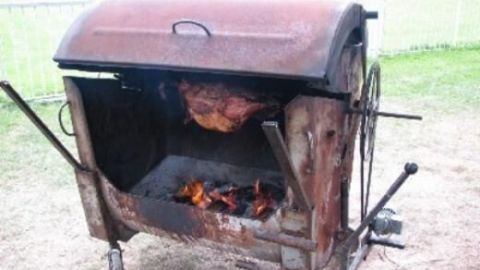 Ein robuster Grill rostet leicht, während Fleisch über offenem Feuer brät; auf einer Rasenfläche im Freien.