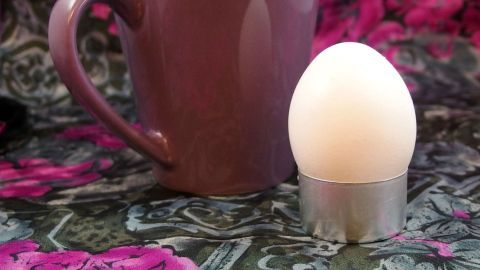 Aluminium-Hüllen von Teelichtern als Eierbecher-Ersatz verwenden. Ein Eierbecher steht neben einer Tasse auf einem gemusterten Tischcloth, umgeben von bunten Blumen.