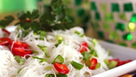 Ein frischer Asia-Nudelsalat mit Glasnudeln, Tomaten und frischen Kräutern auf einem weißen Teller in einem hellen Umfeld.
