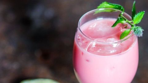 Ein rosa Cocktail steht in einem Glaskelch mit Eis, umgeben von Limetten und grünen Blättern auf einer rustikalen Oberfläche.