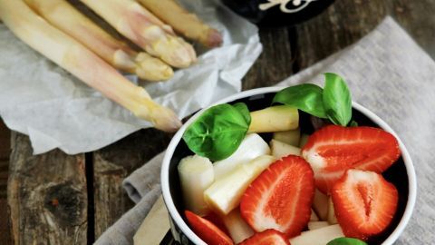 Ein frischer Spargelsalat mit Erdbeeren und Basilikum ist in einer schwarzen Schüssel angerichtet, umgeben von frischem Spargel.