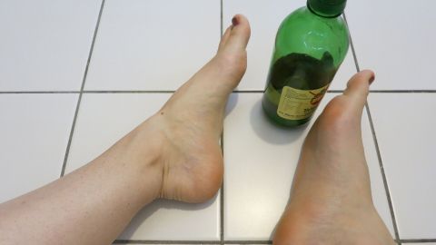 Zwei Füße stehen barfuß auf einem weißen Fliesenboden, neben einer grünen Flasche für ein Essigbad gegen Fußpilz.