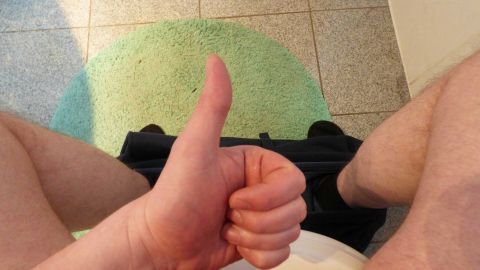 Eine Person sitzt auf der Toilette und zeigt mit Daumen nach oben. Der Boden ist gefliest und ein Teppich liegt darunter.