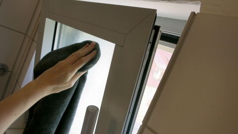 Mit dieser Mischung aus Wasser und etwas Seife kann man Einiges im Haushalt sparsam und effektiv putzen. Eine Hand reinigt ein Fenster mit einem Tuch, während es in einem hellen Raum steht. Das Fenster hat einen klaren Blick nach draußen.