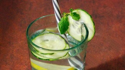 Ein klarer Longdrink mit Gurken- und Zitronenscheiben, serviert mit einem Strohhalm auf einer rustikalen, braunen Oberfläche.