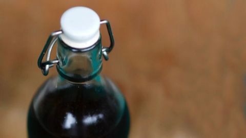 Eine Glasflasche mit Olivenöl steht auf einem Holzuntergrund, während schwarze Oliven daneben liegen, umgeben von einem natürlichen Ambiente.