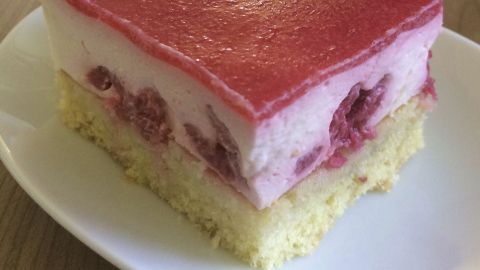 Ein Stück Himbeer-Sauerrahmschnitten liegt auf einem weißen Teller, glänzt rot und enthält eine saftige Schicht Himbeeren.