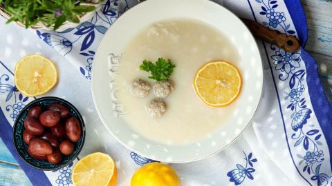 Ein Teller mit Jouvarlakia zeigt die Hackbällchen in einer hellen Suppe, umgeben von Zitronen und einer bunten Tischdecke.