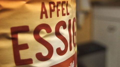Apfelessig hilft sehr gut gegen Halsschmerzen ... Da kann die Erkältung ja kommen. Eine Flasche Apfelessig steht im Vordergrund, während der Hintergrund unscharf eine Küche zeigt.