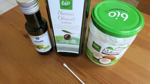 Hilfe bei Schnupfen: Öl mit einem Wattestäbchen direkt in die Nasenlöcher einreiben! Eine Flasche mit Öl, eine Dose mit einer Creme und ein Wattestäbchen liegen auf einem Holztisch, bereit zur Anwendung gegen Schnupfen.