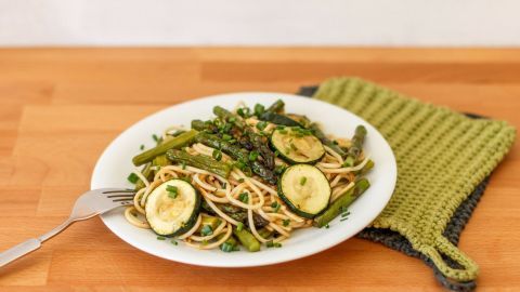 Eine weiße Platte mit Pasta, grünem Spargel und Zucchini, liegt auf einem Holzuntergrund, umgeben von einem grünen Topflappen.